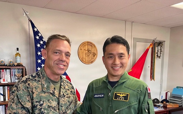 Two military personnel are shaking hands in what appears to be a military office setting. One individual is wearing U.S. Marine Corps digital camouflage (MARPAT) uniform with a name tape reading 'DETRINIS', while the other is wearing a green Japanese military flight suit with 'JAPAN' and 'T. MAEDA' name tape visible. The setting suggests an official bilateral military meeting or exchange visit.