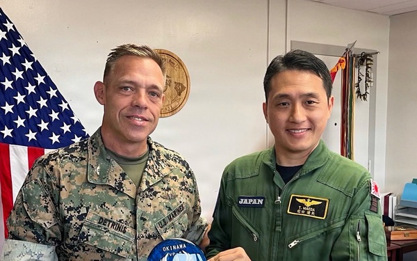 Two military personnel — a U.S. Marine in MARPAT digital camouflage and a Japan Air Self-Defense Force officer in a green flight suit — pose together in an office setting. They are jointly holding a commemorative patch or coin reading 'OKINAWA' with a bird emblem, suggesting a bilateral military exchange or courtesy visit. The setting includes a U.S. flag, an official seal, and a desk consistent with a command office.