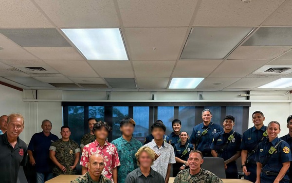 A group photo taken in a conference room showing military personnel in camouflage uniforms, first responders in navy blue uniforms, civilians, and several minors whose faces have been blurred for privacy. Two military personnel in Marine Corps MARPAT camouflage kneel in the front alongside a child, while the larger group stands behind a conference table. The setting appears to be an official meeting or recognition event.