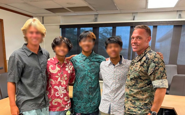 Five individuals are posing for a group photo in what appears to be a conference or meeting room. The group includes one person in U.S. Marine Corps MARPAT digital camouflage uniform on the right, one adult in civilian clothing on the left, and three younger individuals in the center wearing casual button-up shirts. The setting appears to be an official or institutional indoor environment.