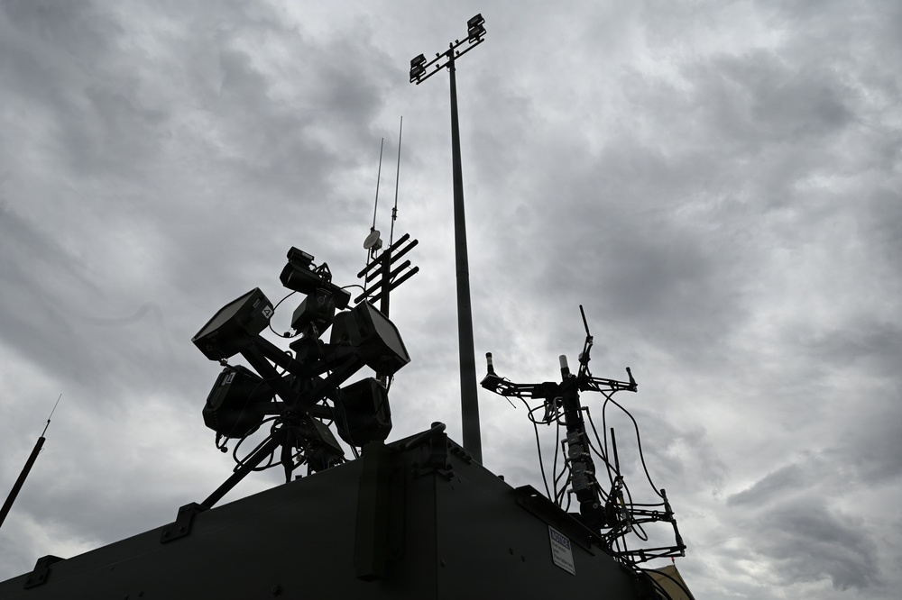 The image shows a silhouetted rooftop or vehicle-mounted communications and surveillance equipment cluster photographed from a low angle against an overcast sky. Multiple antennas, sensor arrays, cameras, and electronic equipment are visible in dark silhouette. The dramatic cloudy sky provides a high-contrast backdrop that obscures fine equipment details.