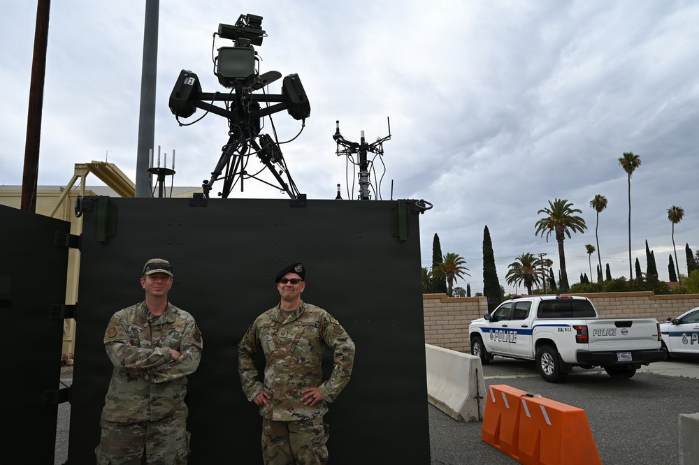 Two military personnel in OCP (Operational Camouflage Pattern) uniforms stand in front of a large black equipment enclosure topped with what appears to be a counter-drone or surveillance system on a tripod mount. The setting appears to be a secured outdoor area, likely a military or government installation, with police vehicles visible in the background. Palm trees and a cloudy sky are visible in the background, suggesting a Southern California or similar warm-climate location.