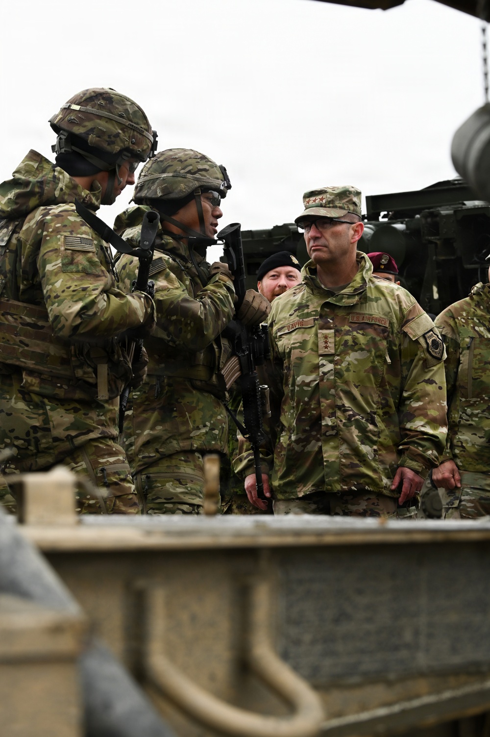 A group of U.S. military personnel in OCP (Operational Camouflage Pattern) uniforms are gathered in what appears to be a field training or inspection environment. A senior U.S. Air Force officer with the name tape 'DAVIS' and four-star rank insignia on his patrol cap is observing soldiers who are handling rifles. Military vehicles and equipment are visible in the background.