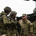 A group of U.S. military personnel in OCP (Operational Camouflage Pattern) uniforms are gathered in what appears to be a field training or inspection environment. A senior U.S. Air Force officer with the name tape 'DAVIS' and four-star rank insignia on his patrol cap is observing soldiers who are handling rifles. Military vehicles and equipment are visible in the background.