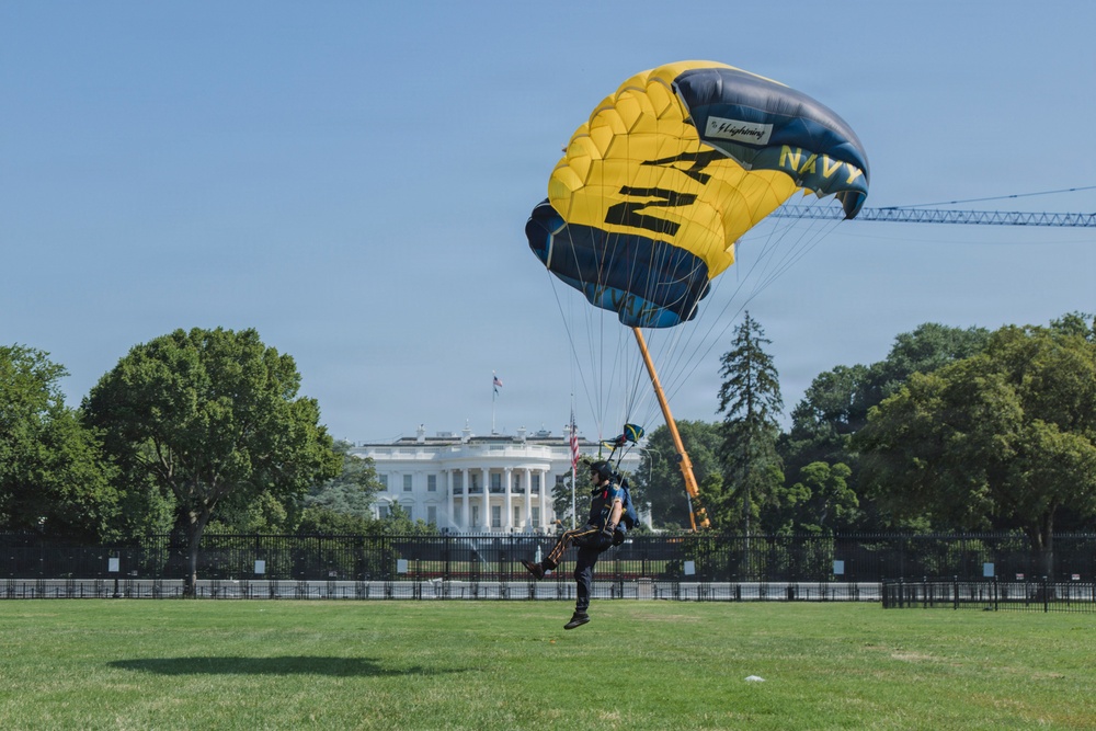 A U.S. Navy parachutist is landing on the Ellipse lawn in front of the White House in Washington, D.C. The parachute is prominently marked with 'NAVY' in blue and yellow colors. This appears to be a demonstration jump, likely part of a military or patriotic event.