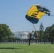 A U.S. Navy parachutist is landing on the Ellipse lawn in front of the White House in Washington, D.C. The parachute is prominently marked with 'NAVY' in blue and yellow colors. This appears to be a demonstration jump, likely part of a military or patriotic event.
