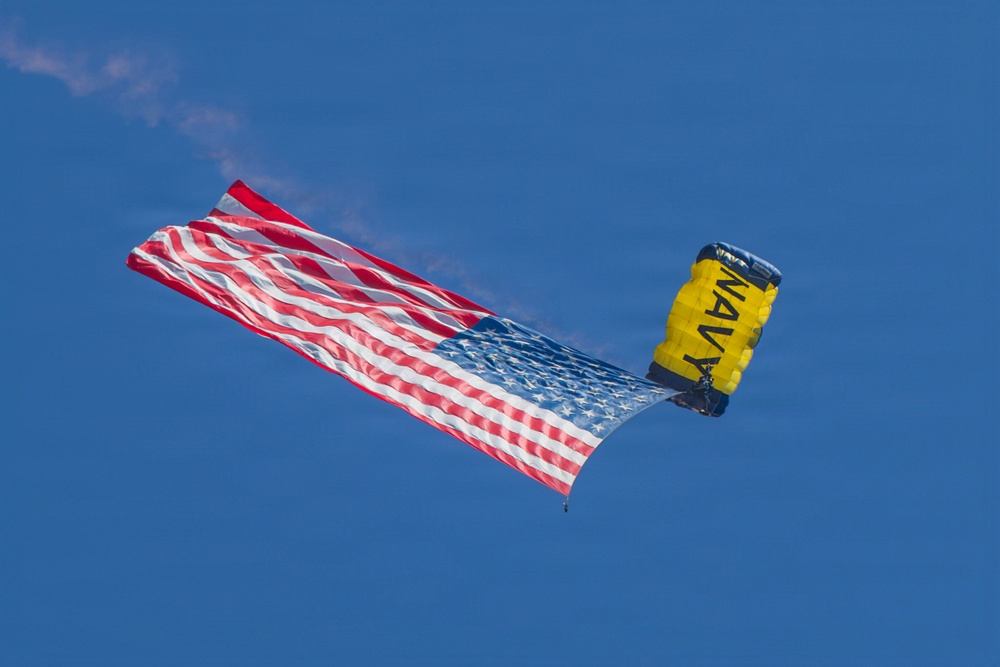 A U.S. Navy parachutist is descending with a large American flag trailing behind them against a clear blue sky. The parachute is distinctively colored in yellow and dark blue with 'NAVY' printed on it. A white smoke trail is visible, suggesting this is part of a ceremonial or airshow demonstration.