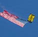 A U.S. Navy parachutist is descending with a large American flag trailing behind them against a clear blue sky. The parachute is distinctively colored in yellow and dark blue with 'NAVY' printed on it. A white smoke trail is visible, suggesting this is part of a ceremonial or airshow demonstration.