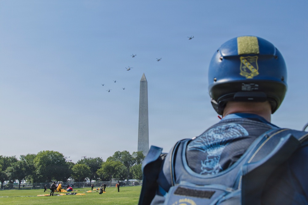A military parachutist in blue gear observes a formation of helicopters flying past the Washington Monument on the National Mall in Washington, D.C. The individual is wearing a blue helmet with a unit patch and a blue jumpsuit with an eagle/parachutist insignia patch on the back. The scene appears to be part of a military demonstration or ceremonial flyover event.