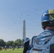 A military parachutist in blue gear observes a formation of helicopters flying past the Washington Monument on the National Mall in Washington, D.C. The individual is wearing a blue helmet with a unit patch and a blue jumpsuit with an eagle/parachutist insignia patch on the back. The scene appears to be part of a military demonstration or ceremonial flyover event.
