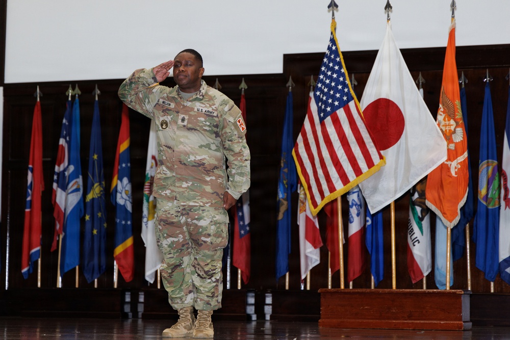 A U.S. Army soldier in OCP (Operational Camouflage Pattern) uniform is rendering a salute during what appears to be a formal military ceremony. The soldier's name tape reads 'MELVIN' and the uniform displays 'U.S. ARMY' tape along with various patches. The setting includes a display of multiple flags including the American flag and the Japanese flag, suggesting a joint or international military event.