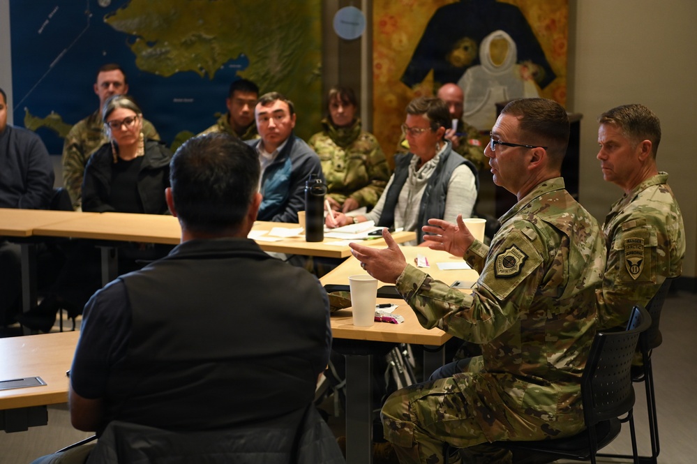 A military briefing or meeting is taking place in what appears to be a conference or community room. Two uniformed military personnel in OCP (Operational Camouflage Pattern) uniforms are seated at a table, with one actively speaking and gesturing with his hands. They are addressing a mixed group of military and civilian attendees seated across from them.