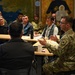 A military briefing or meeting is taking place in what appears to be a conference or community room. Two uniformed military personnel in OCP (Operational Camouflage Pattern) uniforms are seated at a table, with one actively speaking and gesturing with his hands. They are addressing a mixed group of military and civilian attendees seated across from them.