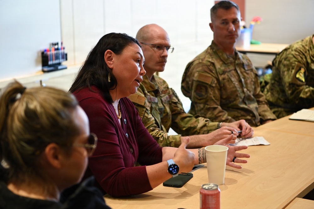 A meeting or briefing is taking place at a table where a civilian woman in a maroon cardigan appears to be speaking or presenting to a group that includes at least three military personnel in camouflage uniforms. The setting appears to be an indoor conference or meeting room. The interaction suggests a formal discussion or community engagement session between civilian and military participants.
