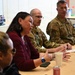 A meeting or briefing is taking place at a table where a civilian woman in a maroon cardigan appears to be speaking or presenting to a group that includes at least three military personnel in camouflage uniforms. The setting appears to be an indoor conference or meeting room. The interaction suggests a formal discussion or community engagement session between civilian and military participants.