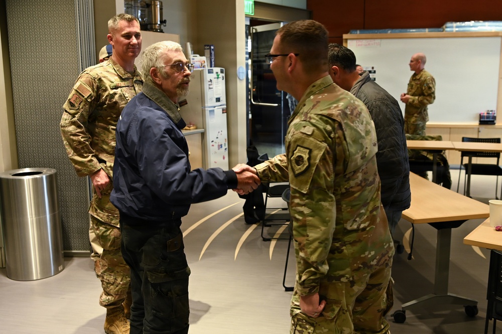 A civilian man in a blue jacket is shaking hands with a military service member in OCP (Operational Camouflage Pattern) uniform in what appears to be a break room or common area of a military facility. Several other uniformed personnel and civilians are present in the background. The setting suggests an informal meeting, recognition event, or visit.