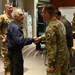 A civilian man in a blue jacket is shaking hands with a military service member in OCP (Operational Camouflage Pattern) uniform in what appears to be a break room or common area of a military facility. Several other uniformed personnel and civilians are present in the background. The setting suggests an informal meeting, recognition event, or visit.