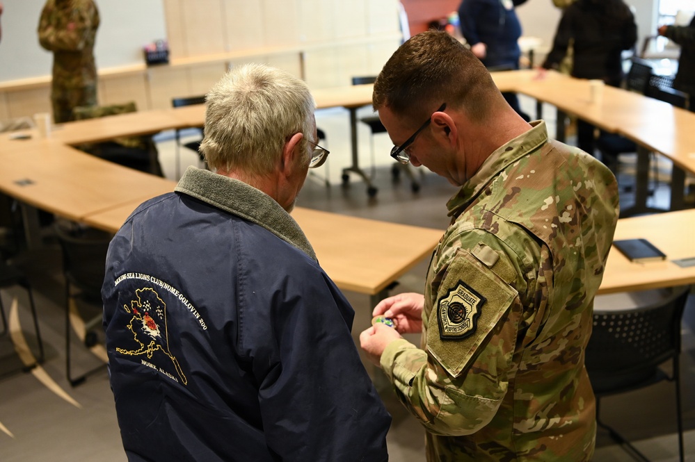 A military service member in MultiCam/OCP camouflage uniform is presenting or pinning something to a civilian wearing a navy blue jacket. The interaction takes place in what appears to be a conference or briefing room with U-shaped table arrangement. The civilian's jacket bears text referencing 'Bering Sea Lions Club / Nome-Golovin' and 'Nome, Alaska.'
