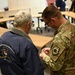 A military service member in MultiCam/OCP camouflage uniform is presenting or pinning something to a civilian wearing a navy blue jacket. The interaction takes place in what appears to be a conference or briefing room with U-shaped table arrangement. The civilian's jacket bears text referencing 'Bering Sea Lions Club / Nome-Golovin' and 'Nome, Alaska.'