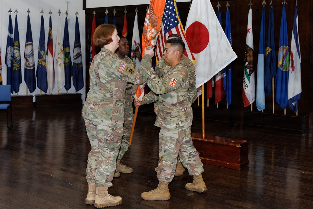 A military change of command ceremony is taking place indoors, with two U.S. Army soldiers in OCP (Operational Camouflage Pattern) uniforms exchanging a unit guidon (orange flag). A third soldier stands at attention in the background. The ceremony is set in a formal hall decorated with numerous U.S. state flags, the American flag, and the Japanese national flag.