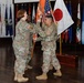 A military change of command ceremony is taking place indoors, with two U.S. Army soldiers in OCP (Operational Camouflage Pattern) uniforms exchanging a unit guidon (orange flag). A third soldier stands at attention in the background. The ceremony is set in a formal hall decorated with numerous U.S. state flags, the American flag, and the Japanese national flag.