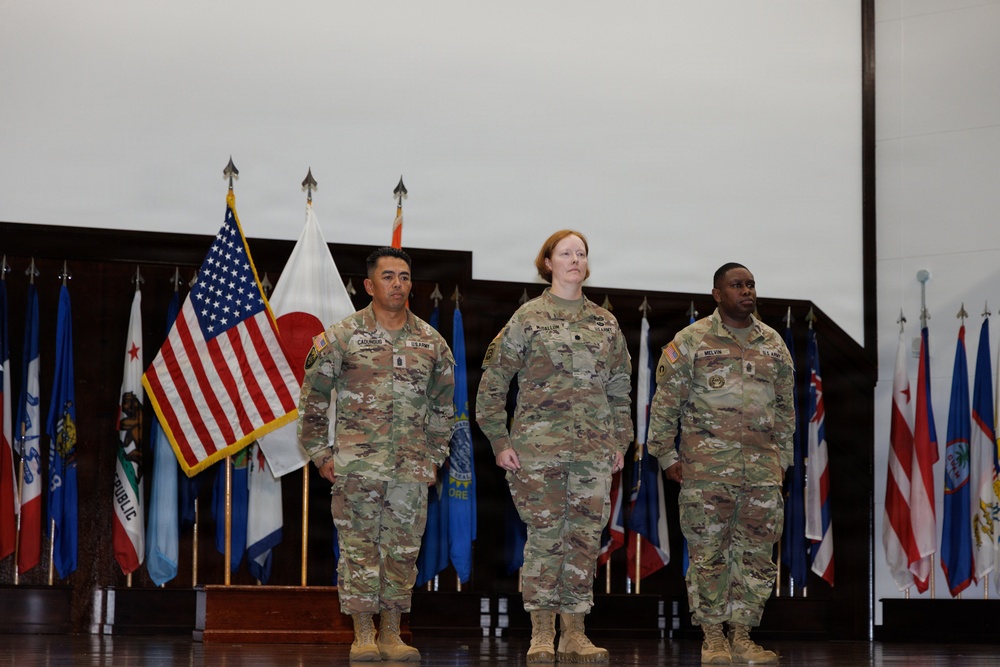 Three U.S. Army soldiers in OCP (Operational Camouflage Pattern) uniforms stand at attention on a stage during a formal military ceremony. They are positioned in front of a display of multiple flags including the U.S. flag and various state and territorial flags. Name tapes visible on the uniforms read 'CASUNDO,' 'DHALLION' (or similar), and 'MELVIN.'