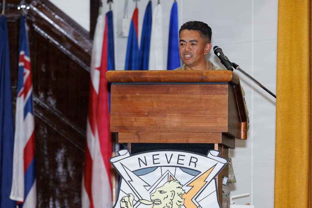 A military service member in uniform is speaking at a wooden podium equipped with a microphone. The podium displays a unit insignia/crest featuring the word 'NEVER,' a bison, and a lightning bolt. Multiple flags are visible in the background, suggesting an official military ceremony or event.