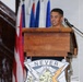 A military service member in uniform is speaking at a wooden podium equipped with a microphone. The podium displays a unit insignia/crest featuring the word 'NEVER,' a bison, and a lightning bolt. Multiple flags are visible in the background, suggesting an official military ceremony or event.