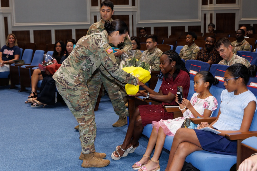 A U.S. Army soldier in OCP (Operational Camouflage Pattern) uniform is presenting a yellow flower bouquet to a seated civilian woman during what appears to be a formal military ceremony in an auditorium. The woman is accompanied by two young girls, and the audience includes both uniformed military personnel and civilian family members. The setting suggests a recognition, promotion, or change-of-command ceremony.