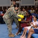 A U.S. Army soldier in OCP (Operational Camouflage Pattern) uniform is presenting a yellow flower bouquet to a seated civilian woman during what appears to be a formal military ceremony in an auditorium. The woman is accompanied by two young girls, and the audience includes both uniformed military personnel and civilian family members. The setting suggests a recognition, promotion, or change-of-command ceremony.