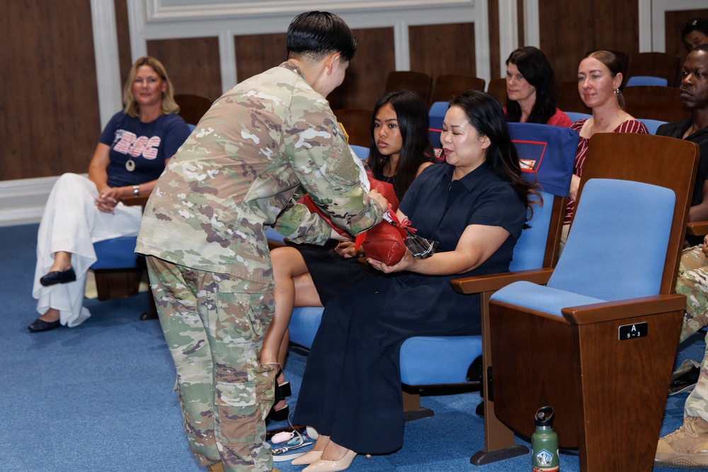 A military ceremony taking place in what appears to be an auditorium or formal meeting room. A soldier in Army Combat Uniform (ACU/OCP camouflage) is presenting a red item, possibly a folded flag or gift, to a seated woman in a black dress, while a younger woman sits beside her. Several other attendees are seated in blue auditorium-style chairs observing the event.