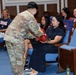 A military ceremony taking place in what appears to be an auditorium or formal meeting room. A soldier in Army Combat Uniform (ACU/OCP camouflage) is presenting a red item, possibly a folded flag or gift, to a seated woman in a black dress, while a younger woman sits beside her. Several other attendees are seated in blue auditorium-style chairs observing the event.