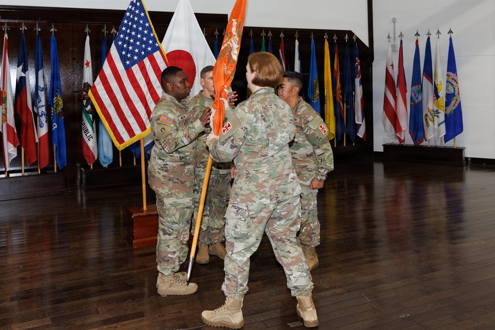 A U.S. Army change of command or activation ceremony is taking place indoors, with soldiers in OCP (Operational Camouflage Pattern) uniforms passing an orange unit guidon. The ceremony is conducted in a formal hall decorated with multiple U.S. state and territory flags along the walls. The passing of the guidon is the central ceremonial act visible in the image.