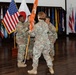 A U.S. Army change of command or activation ceremony is taking place indoors, with soldiers in OCP (Operational Camouflage Pattern) uniforms passing an orange unit guidon. The ceremony is conducted in a formal hall decorated with multiple U.S. state and territory flags along the walls. The passing of the guidon is the central ceremonial act visible in the image.