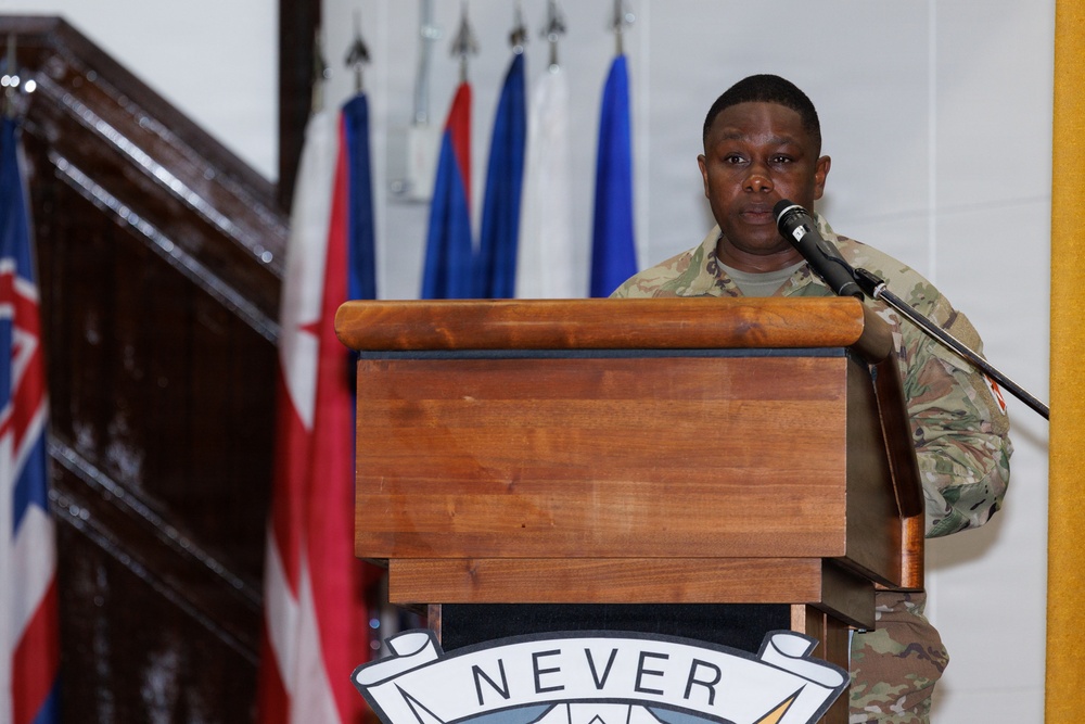 A military service member in MultiCam/OCP camouflage uniform is speaking at a wooden podium equipped with a microphone. The podium displays a partial motto reading 'NEVER' along with what appears to be a unit crest or insignia. Multiple flags are visible in the background, including what appear to be national and unit flags.