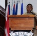 A military service member in MultiCam/OCP camouflage uniform is speaking at a wooden podium equipped with a microphone. The podium displays a partial motto reading 'NEVER' along with what appears to be a unit crest or insignia. Multiple flags are visible in the background, including what appear to be national and unit flags.