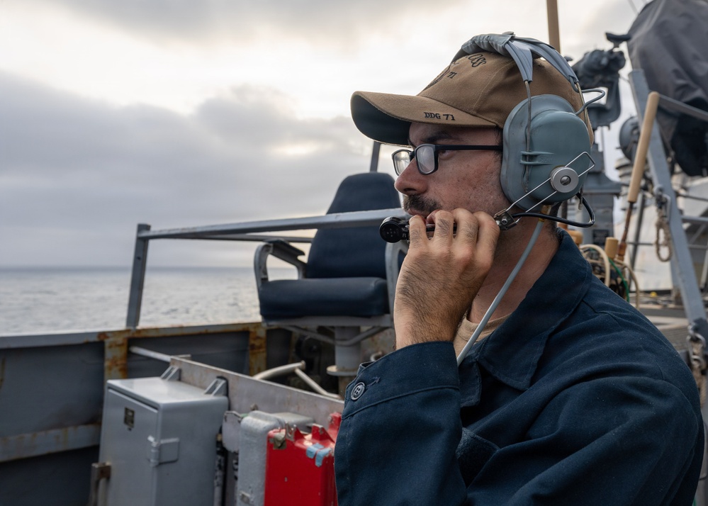 A U.S. Navy sailor is stationed on the deck of a naval vessel, wearing a headset and speaking into a communication device. His cap is marked 'DDG 71', identifying the ship as USS Ross, an Arleigh Burke-class guided-missile destroyer. The sailor is on watch duty, overlooking the open ocean under overcast skies.