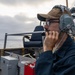 A U.S. Navy sailor is stationed on the deck of a naval vessel, wearing a headset and speaking into a communication device. His cap is marked 'DDG 71', identifying the ship as USS Ross, an Arleigh Burke-class guided-missile destroyer. The sailor is on watch duty, overlooking the open ocean under overcast skies.