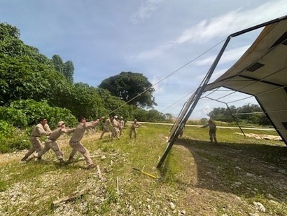 A group of military personnel in tan/desert camouflage uniforms are collectively pulling on ropes attached to a large metal-framed structure, appearing to erect or dismantle a large tent or shelter. The activity takes place in an outdoor area with grassy and rocky terrain surrounded by tropical vegetation. One individual stands near the structure while the others form a line pulling the guide ropes.