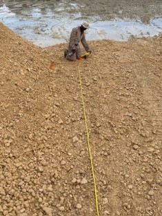 A person in camouflage clothing and a hat is kneeling on a sandy/gravelly earthen berm or embankment, handling what appears to be a yellow measuring tape or cord. The yellow line extends from the person's position down the length of the slope toward the camera. A body of water or wet surface is visible in the background behind the berm.