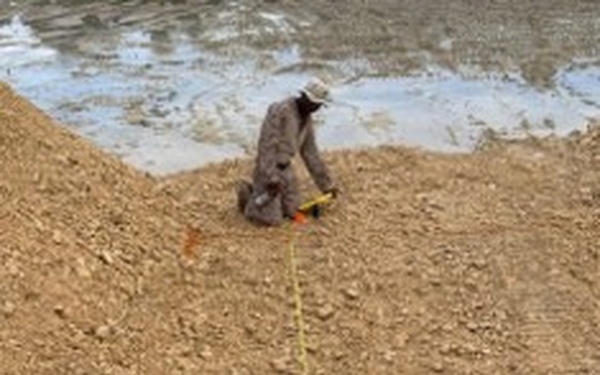 A person in camouflage clothing and a hat is kneeling on a sandy/gravelly earthen berm or embankment, handling what appears to be a yellow measuring tape or cord. The yellow line extends from the person's position down the length of the slope toward the camera. A body of water or wet surface is visible in the background behind the berm.