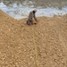 A person in camouflage clothing and a hat is kneeling on a sandy/gravelly earthen berm or embankment, handling what appears to be a yellow measuring tape or cord. The yellow line extends from the person's position down the length of the slope toward the camera. A body of water or wet surface is visible in the background behind the berm.