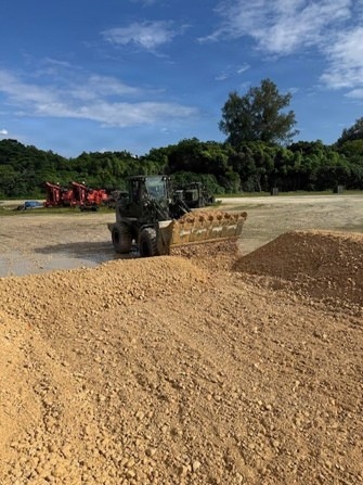 An outdoor construction or earthmoving site showing a wheel loader actively pushing or moving piles of sandy/granular material in the foreground. Multiple pieces of heavy equipment, including red machinery, are visible in the background against a treeline. The scene appears to be a staging or grading area under clear, partly cloudy skies.