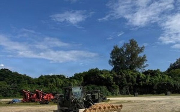 An outdoor construction or earthmoving site showing a wheel loader actively pushing or moving piles of sandy/granular material in the foreground. Multiple pieces of heavy equipment, including red machinery, are visible in the background against a treeline. The scene appears to be a staging or grading area under clear, partly cloudy skies.