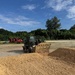 An outdoor construction or earthmoving site showing a wheel loader actively pushing or moving piles of sandy/granular material in the foreground. Multiple pieces of heavy equipment, including red machinery, are visible in the background against a treeline. The scene appears to be a staging or grading area under clear, partly cloudy skies.