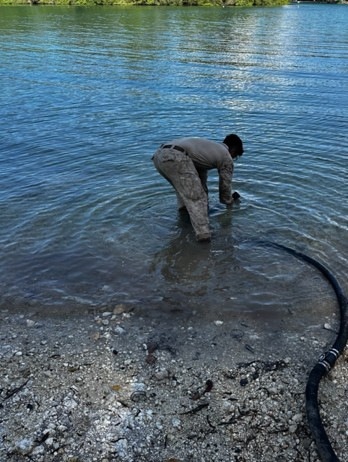 A person wearing camouflage or military-style clothing is bent over in shallow water at the edge of a river or lake, appearing to handle something at the water's surface. A large black hose or pipe extends from the shoreline into the water near the person. The background shows calm blue water with green vegetation along the far bank.