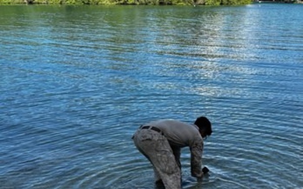 A person wearing camouflage or military-style clothing is bent over in shallow water at the edge of a river or lake, appearing to handle something at the water's surface. A large black hose or pipe extends from the shoreline into the water near the person. The background shows calm blue water with green vegetation along the far bank.