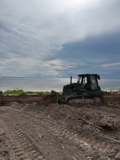 A dark-colored tracked bulldozer is actively grading or clearing land near a coastal shoreline. The foreground shows heavily disturbed, churned soil with visible track marks and debris. The background reveals a calm body of water under a partly cloudy sky.