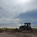 A dark-colored tracked bulldozer is actively grading or clearing land near a coastal shoreline. The foreground shows heavily disturbed, churned soil with visible track marks and debris. The background reveals a calm body of water under a partly cloudy sky.