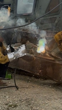 A worker is performing welding or cutting operations on a large piece of heavy metal equipment or machinery, likely outdoors or in an industrial yard. Bright arc light, colorful flame, smoke, and sparks are visible at the work point. The worker is wearing protective leather gloves and appears to be holding a metal component against the workpiece.